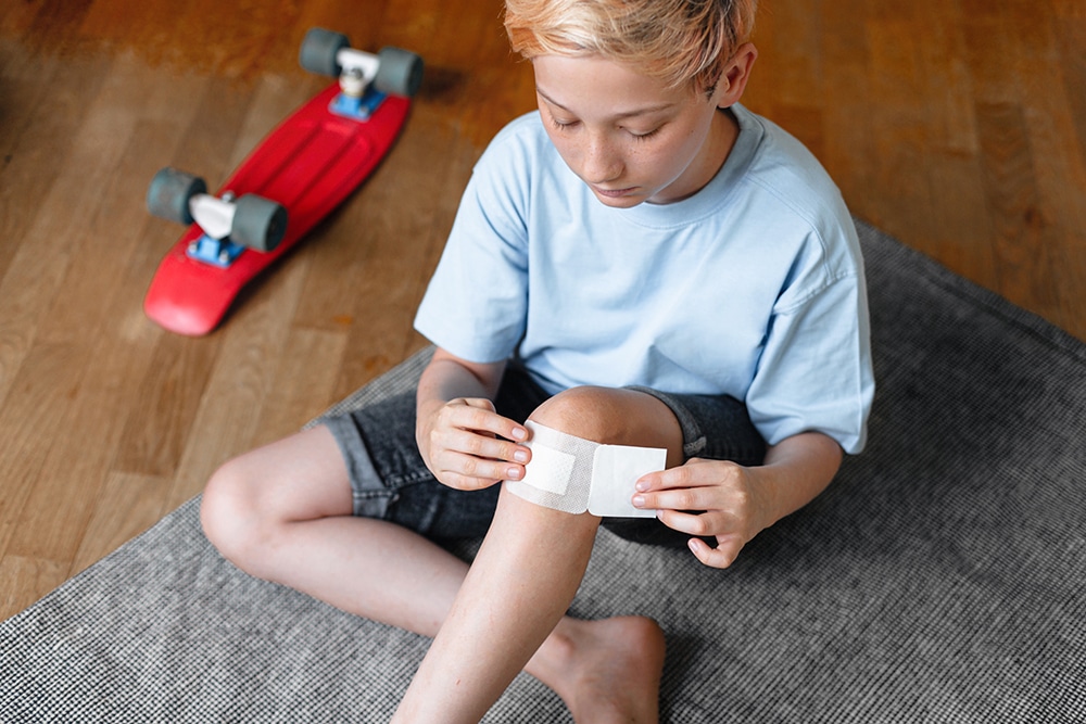 Boy putting sticking plaster on injured knee skin by himself. First aid for wounds.